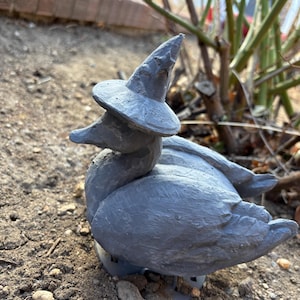May include: A gray duck statue wearing a witch hat, resting on the ground. The duck is painted in a matte gray finish, with visible texture. The witch hat is pointed and has a wide brim. The statue is a whimsical garden decoration.