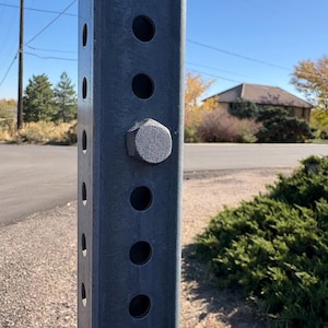 May include: A close-up of a gray metal post with evenly spaced holes and a large bolt. The post is outdoors with a clear blue sky and a house in the background. The post is likely part of a sign or structure.