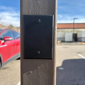 May include: A black rectangular plate with two screws attached to a brown post. The plate is mounted on a weathered, textured post. The background shows a red car, a building, and a blue sky.