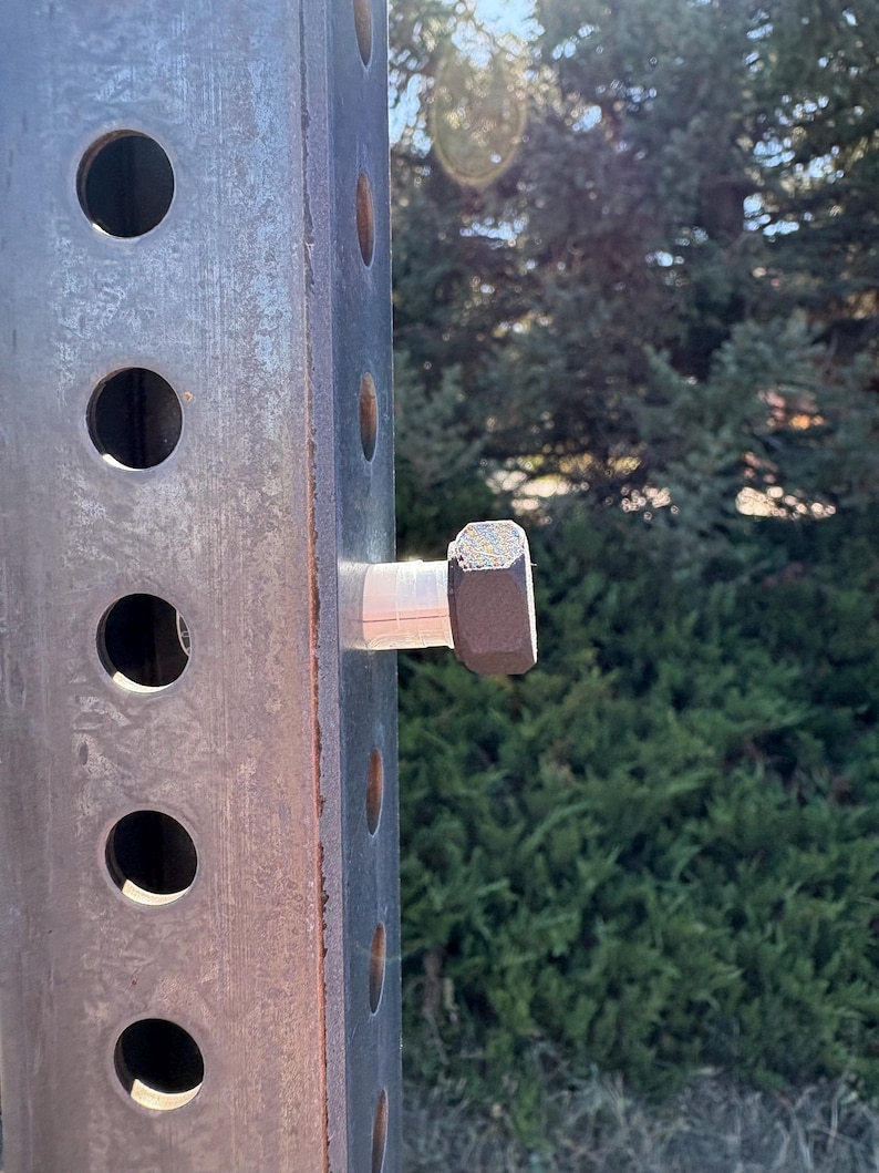 Peut inclure: Gros plan sur un poteau m&eacute;tallique rectangulaire et us&eacute;, perc&eacute; de trous circulaires espac&eacute;s. Un gros boulon hexagonal est fix&eacute; au poteau avec une entretoise blanche. Le poteau a un aspect rouill&eacute;, sugg&eacute;rant une utilisation en ext&eacute;rieur.