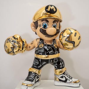 Puede incluir: Estatua dorada de un personaje de dibujos animados con atuendo de boxeo. La figura lleva un sombrero dorado, guantes de boxeo con la palabra "MONEY" y ropa negra y dorada. El personaje tiene tatuajes y posa en posición de combate.