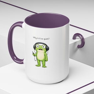 May include: White ceramic mug with a purple interior and handle. The mug features a cartoon frog wearing headphones and holding a small device. Text on the mug reads, "Why is it so quiet?"