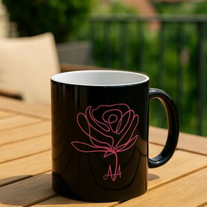 May include: Black ceramic mug featuring a pink line drawing of a rose. The mug has a white interior and a black handle. The initials "AA" are printed in pink below the rose design. The mug is sitting on a wooden table.