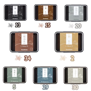 May include: A collection of seven Shachihata ink pads in various colors, each in a rectangular black case. The pads feature Japanese text and decorative floral accents with numbers. Colors include black, brown, gray, and others.