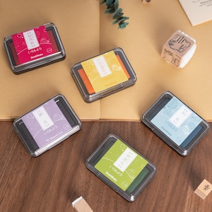May include: A collection of Shachihata ink pads in various colors, including red, orange, purple, green, and blue, each in a clear plastic case. Japanese text is visible on the labels. A small wooden stamp is also present.