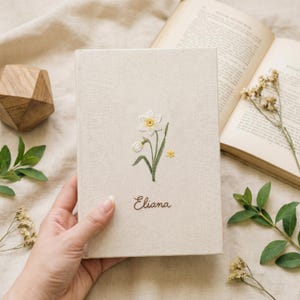 Puede incluir: Un diario beige con un diseño de narciso bordado y el nombre "Eliana" en escritura cursiva. El diario está abierto junto a un libro y un objeto decorativo de madera. La portada presenta un narciso blanco con detalles amarillos y hojas verdes.