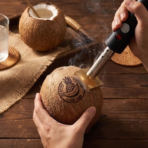 May include: A handheld electric branding iron with a metal heating element and black handle is pressing a circular logo onto a brown coconut. The logo features a pineapple and banana graphic with the text SUN RIPENED SUN FARM.