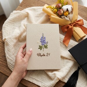 May include: A rectangular, cream-colored fabric-covered journal featuring an embroidered purple flower stalk with green leaves and a small white flower. The text "Chapter 21" is embroidered in brown thread below the floral design on the front cover.