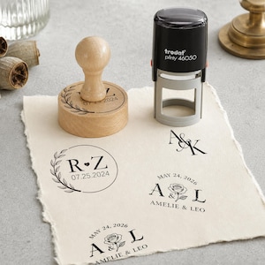 May include: A round wooden handle stamp and a black Trodat Printy 46050 self-inking stamp. The paper displays black ink impressions featuring the initials R&Z with a date, A&K, and A&L with a rose illustration and the names Amelie & Leo.