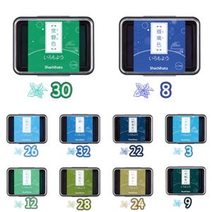 May include: A collection of Shachihata ink pads in various colors, including green, blue, and teal. Each pad is in a rectangular metal case with Japanese text. Numbers and floral illustrations accompany each ink pad.