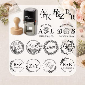 Könnte beinhalten: Ein schwarz-grauer Trodat Printy 46050 Selbstfärbestempel und ein Holzstempel. Das Bild zeigt verschiedene Monogramm-Designs in schwarzer Tinte mit Initialen, Daten, Namen, Blumenkränzen und geometrischen Rändern für individuelle Hochzeitspapeterie.
