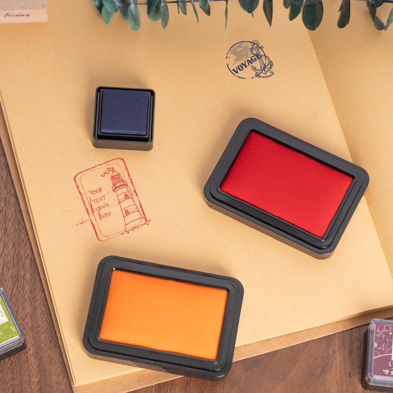 May include: An assortment of ink pads in blue, red, and orange, with black frames, are displayed on a tan paper surface. A blue ink pad is square, while the others are rectangular. A lighthouse stamp with the text "Your text goes here" is visible.