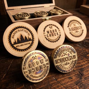 May include: A collection of circular wooden and brass branding irons for woodworking. Three wooden discs feature laser-engraved logos for Stuart Payne, Wood Chuck, and D.G. Couch. Two brass stamps with matching designs sit in front, with a wooden storage box behind.
