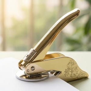May include: A gold-coloured seal press with a floral design on the base. The press is positioned over a white piece of paper, ready for use. The handle is curved and ergonomic, and the overall design is elegant.