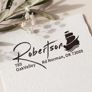 May include: A black ink stamp impression on textured white paper featuring the name Robertson in a cursive script, the address 789 OakValley Rd Norman, OK 73069, and a graphic silhouette of a sailing ship with three distinct waves underneath.