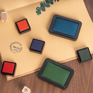 May include: An assortment of ink pads in various colors, including red, blue, green, and purple, with black frames. The image also shows a stamp with the word "PAID" and a small wooden stamp.