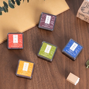 May include: A collection of Shachi-iro ink pads in various colors, including red, orange, green, blue, and purple. Each square pad has Japanese text and a clear plastic lid. The pads are arranged on a wooden surface.