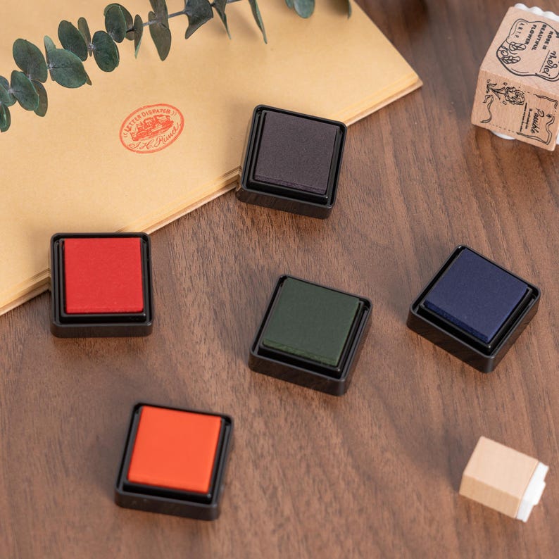 May include: A collection of colorful ink pads in black square cases, including red, orange, green, blue, and brown. A wooden stamp and a brown envelope with a red stamp are also visible on a wooden surface. The ink pads are for crafts and stationery.