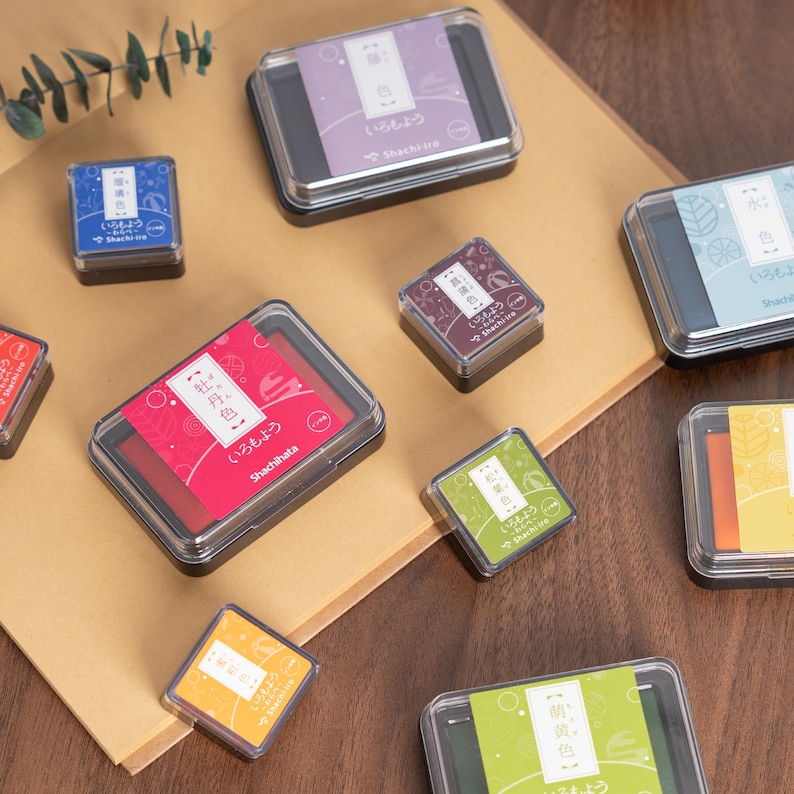 May include: A collection of colorful ink pads in various sizes and colors, including red, purple, green, and yellow. Each pad is in a clear plastic case with Japanese text and designs. The ink pads are arranged on a brown surface.