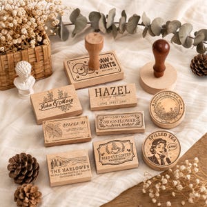 May include: A collection of wooden rubber stamps featuring various designs including John & Mary, Hazel, Moonflower, Bird Cup Coffee, Spilled Tea, and The Harlowes. The stamps have light wood blocks and dark wood handles, displaying intricate black ink illustrations.
