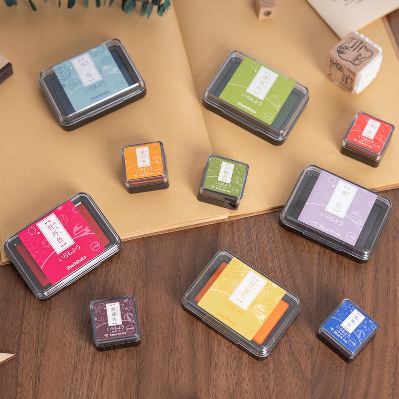 May include: A collection of colorful ink pads in various sizes and shapes. The ink pads are in clear plastic cases with black frames. Colors include blue, green, red, purple, orange, and yellow. Japanese text is visible on the ink pads.