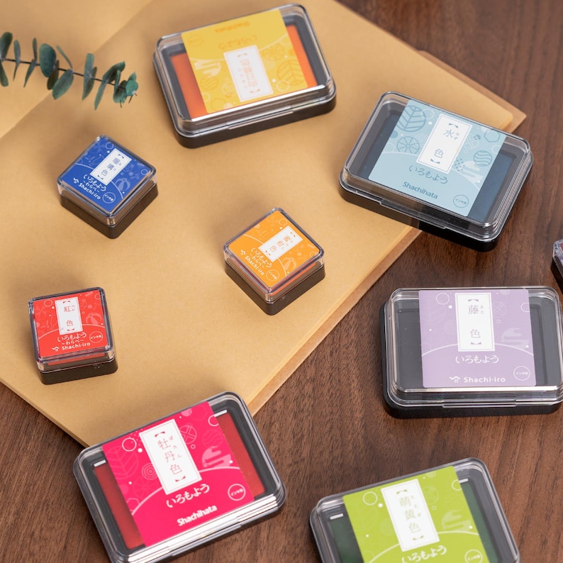 May include: A collection of colorful Shachihata ink pads in various sizes and colors, including red, blue, orange, and green. Each pad is in a clear plastic case with Japanese text. The pads are arranged on a brown surface.