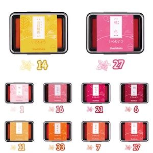 May include: A collection of Shachihata ink pads in various colors, each in a rectangular black case. The pads have labels with Japanese text and decorative elements. Numbers 1, 6, 7, 11, 14, 16, 17, 21, 27, and 33 are visible.