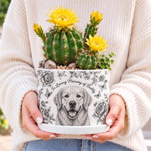May include: A white ceramic planter featuring a black and white illustration of a golden retriever dog surrounded by floral and butterfly motifs. The text "In Loving Memory of Buddy" is printed above the dog. The planter holds green cacti.