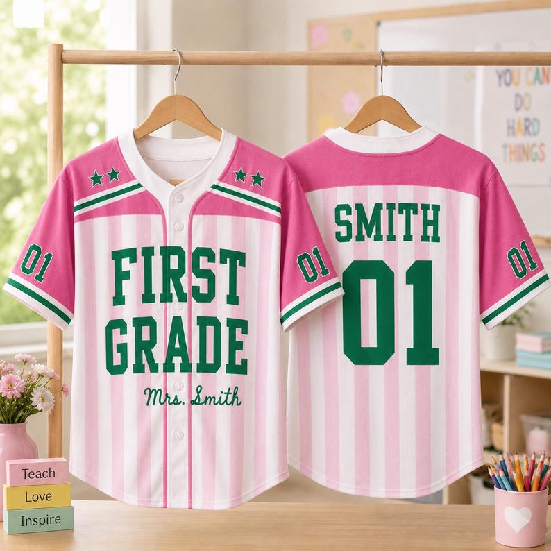 May include: A pink and white vertical striped baseball jersey with green trim. The front features the text FIRST GRADE and Mrs. Smith with two green stars. The back displays the name SMITH and the number 01 in large green block lettering.