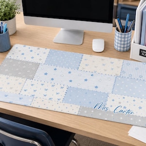 May include: A rectangular desk mat featuring a patchwork design of light blue, white, and gray squares with patterns of stars, bows, and stripes. The bottom right corner displays the name "Mrs. Carter" in a blue cursive script font.