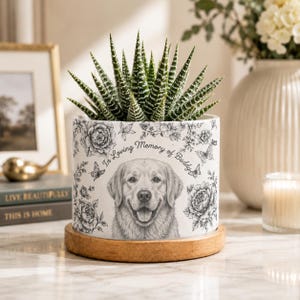 May include: A white ceramic planter with a wooden base containing a green succulent. The planter features a black and white illustration of a golden retriever, floral patterns, butterflies, and the text In Loving Memory of Buddy.