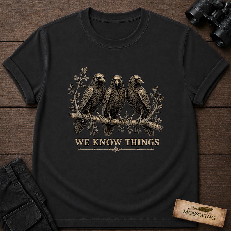 May include: A black short-sleeved t-shirt featuring a graphic of three crows perched on a branch with foliage. Below the birds, the text reads WE KNOW THINGS in a tan color, accompanied by a decorative horizontal line with a small central emblem.
