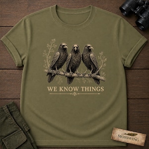 May include: An olive green t-shirt featuring a graphic of three black crows perched on a branch. Below the birds, the text reads WE KNOW THINGS in a light-colored font. The design includes subtle botanical line art surrounding the birds.