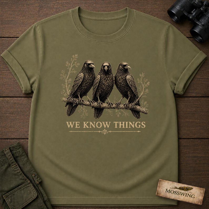 May include: An olive green t-shirt featuring a graphic of three black crows perched on a branch. Below the birds, the text reads WE KNOW THINGS in a light-colored font. The design includes subtle botanical line art surrounding the birds.