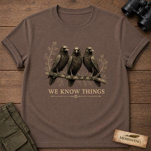 May include: A brown short-sleeved t-shirt featuring a graphic of three black crows perched on a branch with light-colored foliage. Below the birds, the text "WE KNOW THINGS" is printed in a serif font, accompanied by a decorative horizontal line.