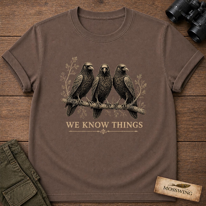 May include: A brown short-sleeved t-shirt featuring a graphic of three black crows perched on a branch with light-colored foliage. Below the birds, the text "WE KNOW THINGS" is printed in a serif font, accompanied by a decorative horizontal line.
