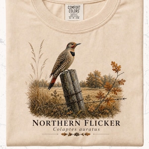 May include: Cream-colored t-shirt featuring a detailed illustration of a Northern Flicker bird perched on a wooden post. The design includes a rural landscape with text that reads "Northern Flicker Colaptes auratus".