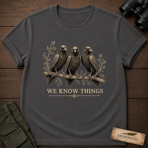 May include: A charcoal gray short-sleeved t-shirt featuring a graphic of three black crows perched on a branch with surrounding foliage. Below the birds, the text WE KNOW THINGS is printed in a light tan color with a decorative line underneath.