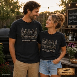 May include: A black t-shirt featuring a graphic print of three crows perched on a branch with the text WE KNOW THINGS printed below the birds. The design is rendered in muted earth tones, showcasing detailed illustrations of the birds and foliage.