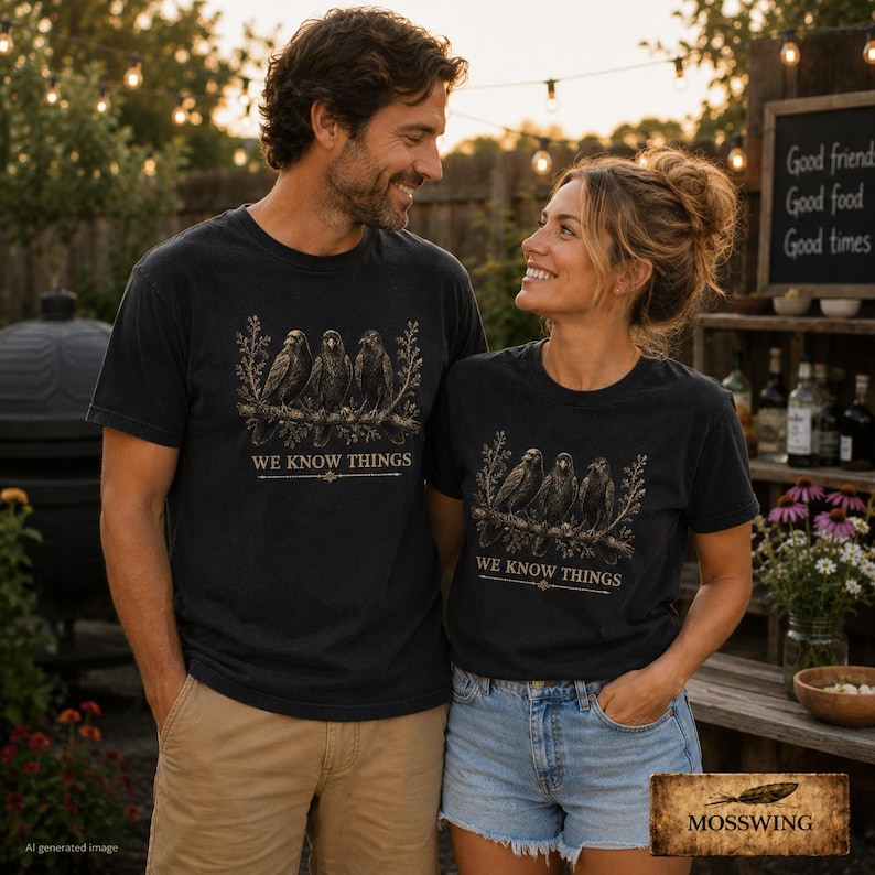 May include: A black t-shirt featuring a graphic print of three crows perched on a branch with the text WE KNOW THINGS printed below the birds. The design is rendered in muted earth tones, showcasing detailed illustrations of the birds and foliage.