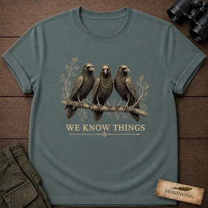 May include: A slate blue short-sleeved t-shirt featuring a graphic of three black crows perched on a branch. The design includes light-colored botanical accents and the text WE KNOW THINGS printed in a serif font below the birds on the center chest.