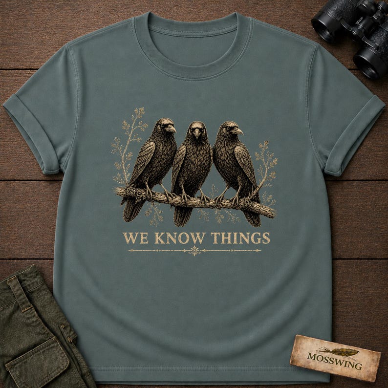 May include: A slate blue short-sleeved t-shirt featuring a graphic of three black crows perched on a branch. The design includes light-colored botanical accents and the text WE KNOW THINGS printed in a serif font below the birds on the center chest.
