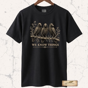 May include: A black short-sleeve t-shirt featuring a graphic of three crows perched on a branch with foliage. Below the birds, the text WE KNOW THINGS is printed in a gold-toned font, accompanied by a decorative horizontal line element.