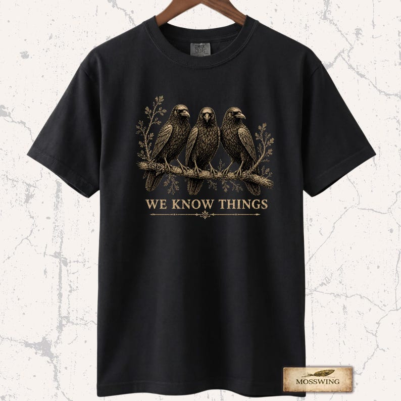 May include: A black short-sleeve t-shirt featuring a graphic of three crows perched on a branch with foliage. Below the birds, the text WE KNOW THINGS is printed in a gold-toned font, accompanied by a decorative horizontal line element.