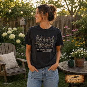 May include: A charcoal gray short-sleeve t-shirt featuring a graphic print of three crows perched on a branch with the text WE KNOW THINGS printed below the birds in a gold-toned font. The shirt is worn with blue denim jeans.