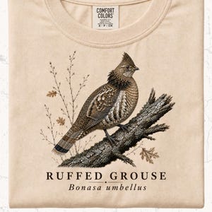 May include: A beige Comfort Colors t-shirt featuring a detailed illustration of a Ruffed Grouse perched on a tree branch. The bird is brown and tan with intricate feather patterns. The text "RUFFED GROUSE Bonasa umbellus" is printed below the image.