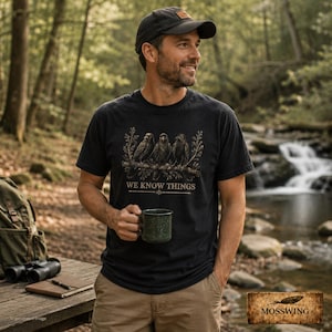 May include: A black short-sleeved t-shirt featuring a graphic of three crows perched on a branch with the text WE KNOW THINGS printed below. The shirt is worn with a dark baseball cap and tan pants, paired with a green backpack and binoculars.