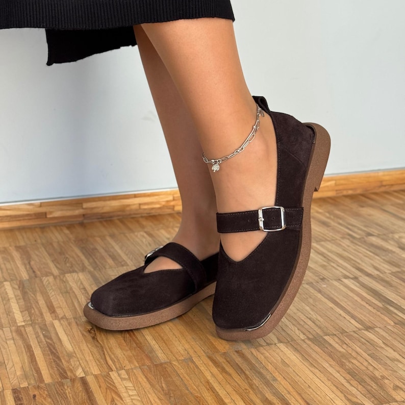 May include: Brown suede Mary Jane style shoes with a buckle closure. The shoes have a rounded toe and a thick, light brown sole. The shoes are worn with a silver anklet.