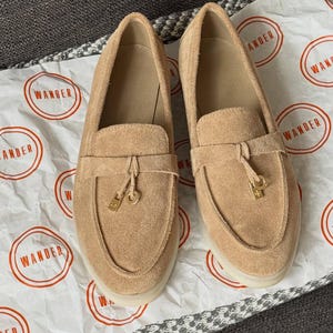 May include: A pair of tan suede loafers featuring a moccasin-style toe, a horizontal strap across the vamp, and a small gold-toned metal charm hanging from the center of each strap. The shoes have a light-colored sole and a smooth interior lining.