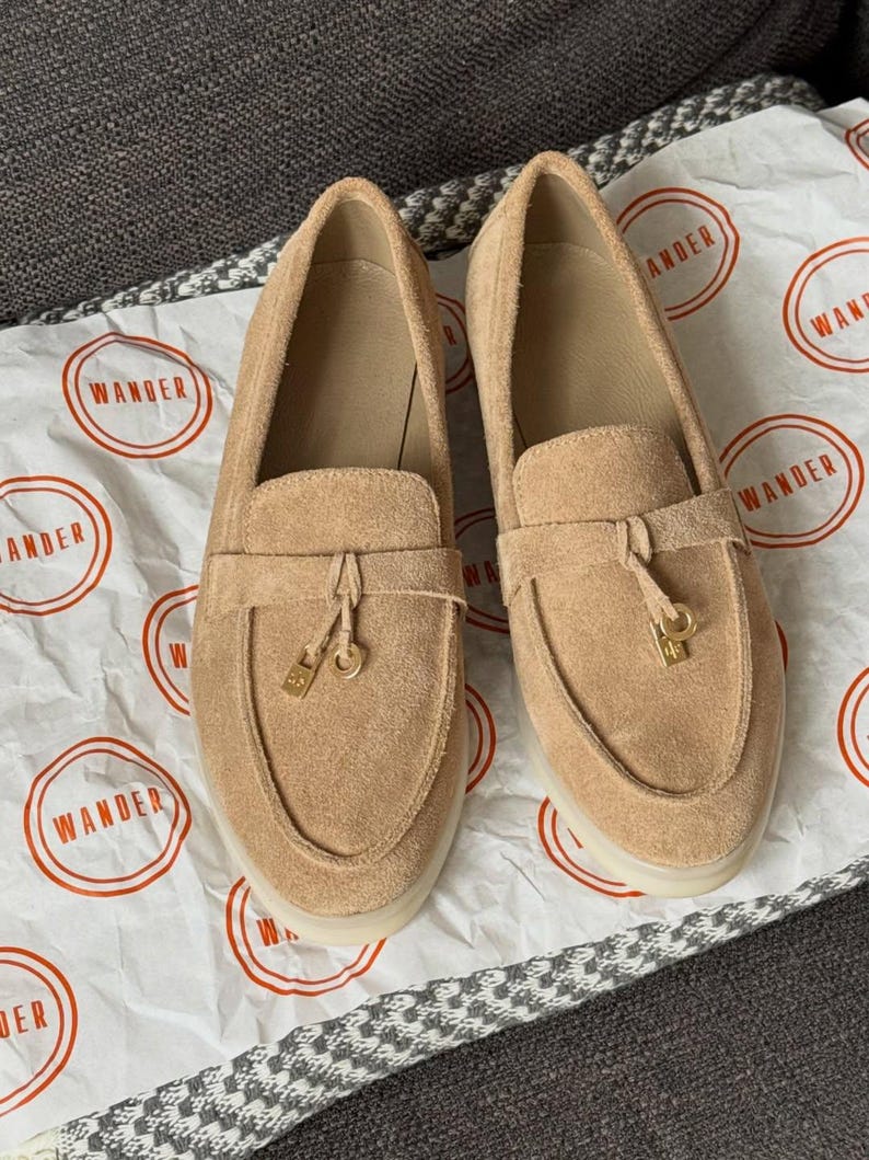 May include: A pair of tan suede loafers featuring a moccasin-style toe, a horizontal strap across the vamp, and a small gold-toned metal charm hanging from the center of each strap. The shoes have a light-colored sole and a smooth interior lining.
